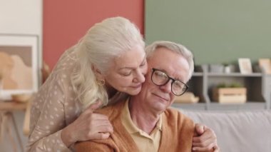 Oturma odasında oturan, beyaz saçlı, üzgün, yaşlı, beyaz gözlüklü bir adamın arkadan sarılıp kocasını güldürmesi.