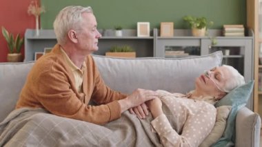 Orta boylu, beyaz saçlı bir adam, kanepede oturan, burnunda oksijen tüpü olan yaşlı bir kadınla yatan, bir kadının elini okşayan bir adam.