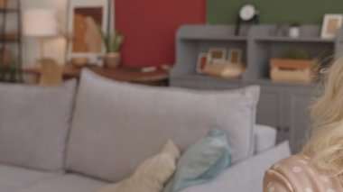 Düşünceli, beyaz saçlı, beyaz bir kadının gündüz vakti oturma odasında oturup mesafeyi izlemesi.