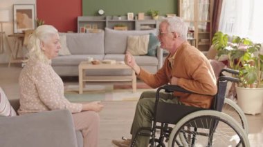 Beyaz saçlı karısının önünde gün ortasında tekerlekli sandalyede oturan beyaz adamın yan görüntüsü. Yaşlı çift taş kağıt makas oynuyor, gülümsüyor ve konuşuyor.