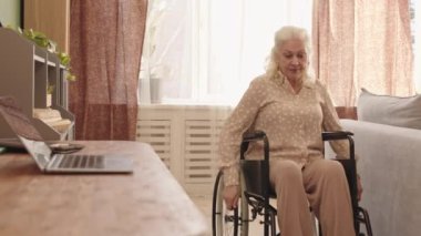 Tekerlekli sandalyedeki yaşlı beyaz kadının odasında masasına yaklaşması ve gündüz vakti taşınabilir bilgisayar kullanması.