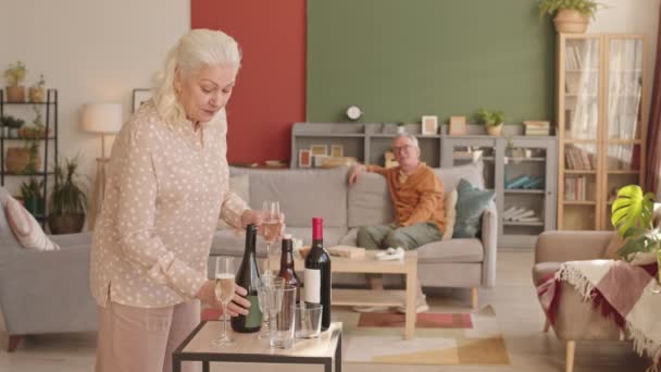 ボトルからワイングラスにスパークリングワインを注ぐ古い白人女性のロックダウン その後 彼女の白い髪の夫にそれらをもたらしますリビングルームでソファに座って — ストック動画