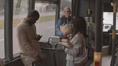 Orta uzunlukta genç siyahi bir adam akıllı telefon ve elinde kitaplarla toplu taşıma aracında dikilip konuşan Asyalı kadın arkadaşını kullanıyor.
