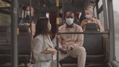 Orta uzunlukta, tıbbi maske ve kulaklık takan, kitap tutan, otobüste duran, tırabzanı tutan, sonra da koltukta oturan siyahi adamla konuşan bir Asyalı kadın.