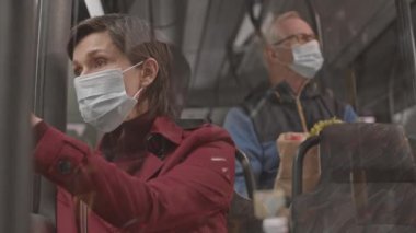 Tıbbi maske takan, kırmızı elbiseli, otobüste oturan, pencereden dışarı bakan olgun, kısa saçlı, beyaz bir kadının göğsü.