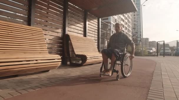 Weiträumige Kamerafahrt eines jungen kaukasischen Mannes im Rollstuhl auf einem Spaziergang im Freien, vorbei an Holzbänken an sonnigen Tagen