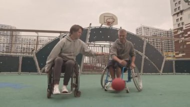 Beyaz kadın ve erkek, tekerlekli sandalyeli, dışarıda basketbol oynayan ve gülümseyen amatör sporcuların geniş açılı çekimi.