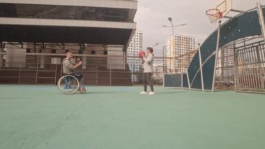 Tekerlekli sandalyedeki beyaz bir adamın önünde duran genç bir kadınla top oynarken görüntüsü, basketbol sahasında gülümseyen insanlar.