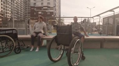 Kadın ve erkek beyaz yetişkinlerin spor sahasında bankta oturup rehabilitasyon egzersizleri yaptığı yaz akşamları bacak kaldırdığı ön planda tekerlekli sandalyeler.