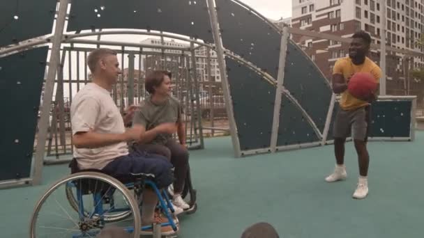 車椅子で白人女性と男性アスリートとバスケットボールをしている認識できない人のPov 屋外コートでそれらを助けるアフリカ系アメリカ人コーチ — ストック動画