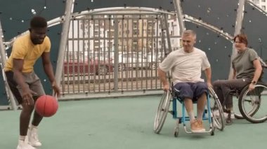 Genç Afrikalı Amerikalı koçla birlikte mahalledeki basketbol sahasında tekerlekli sandalye eğitimi alan gülümseyen beyaz kadın ve erkek arkadaşlar.