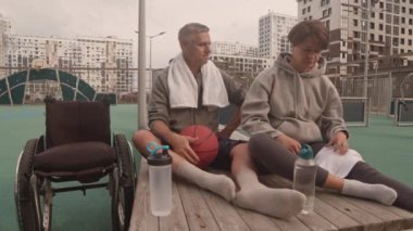 Fiziksel engelli yetişkin beyaz kadın ve erkek sporcuları izliyorlar. Ayakları yukarıda bankta oturuyorlar, antremandan sonra konuşuyorlar. Tekerlekli sandalyeleri yanlarında.
