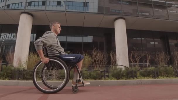 白人年轻男性残疾人坐轮椅 经过多层建筑时的低视角侧视跟踪 — 图库视频影像