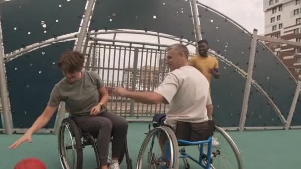 若いアフリカ系アメリカ人バスケットボールコーチの訓練の長いショット白人女性と車椅子で男屋外コートでバスケットボールをプレイ — ストック動画