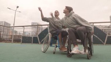 Beyaz kadın ve erkek, tekerlekli sandalyeli, dışarıda basketbol oynayan ve gülümseyen amatör sporcuların düşük açılı görüntüsü.