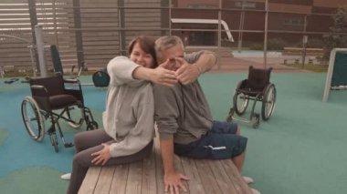 Pozitif beyaz kadın ve fiziksel engelli bir adamın yaz akşamları spor salonunda sırt sırta oturması, arka planda tekerlekli sandalyeler.