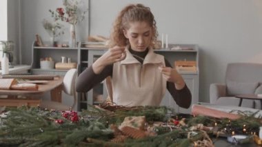 Beyaz tenli genç bir zanaatkar gün ortasında atölyesinde oturuyor ve Noel çelengi yapıyor.