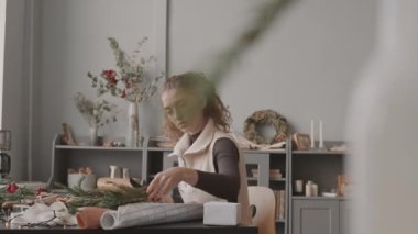 Beyaz tenli genç kadın gündüz atölyesinde masa başında oturuyor, Noel ağacı dallarını makasla kesiyor.