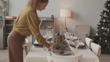 Orta uzunluktaki beyaz bir kadın yemek masasına mum koyuyor. Noel için dekore ediyor, sonra da onları aydınlatmış olanlardan yakıyor.