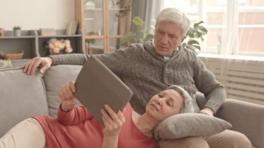 Orta yaşlı, mutlu Asyalı bir kadın. Kafkasyalı kocasının kucağında dinleniyor. Evdeki kanepede oturuyor. Karısı tablet bilgisayar kullanıyor.