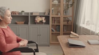 Kıdemli Kafkas kocasının kısa saçlı Asyalı karısını tekerlekli sandalyeye oturttuğu, masasına uğradığı, taşınabilir bilgisayarda daktilo çeken kadının kilitli yan görüntüsü.