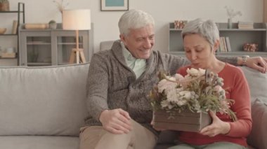 Orta uzunlukta mutlu, kısa saçlı, Asyalı bir kadın. Evdeki kanepede oturuyor. Kafkasyalı kocasının sepetindeki güzel çiçeklere hayran.