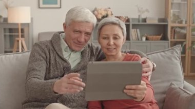 Orta yaşlı, beyaz bir adam elinde tablet bilgisayar olan Asyalı karısına sarılıyor. Evdeki kanepede oturuyor.