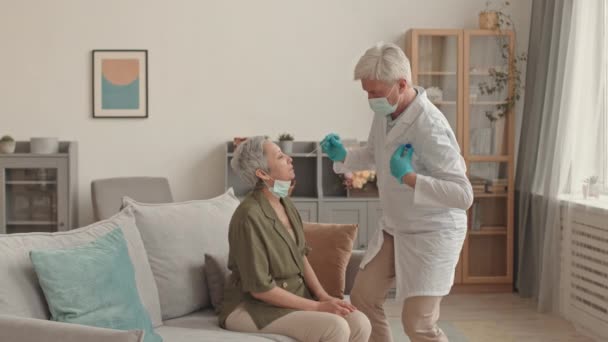 白种人男性男性医生 白天身穿洗涤剂 手套和面罩 用棉签从坐在家中沙发上的亚洲老年妇女的鼻子上进行验血 — 图库视频影像