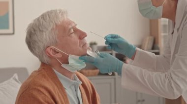 Kadın Asyalı doktor önlük, eldiven ve maske giyer ve Coronavirus testi yaptırır. Kıdemli beyaz adamın burnundaki pamukla gündüz vakti evde oturur.