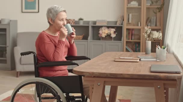 侧视图中等长度的矮个子亚洲老年妇女坐在轮椅上 坐在桌旁 早上喝着杯子里的热茶 望着窗外 — 图库视频影像