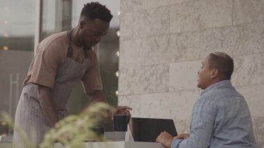 Orta uzunlukta siyah erkek garson, açık havada masada oturan, taşınabilir bilgisayar kullanan genç çift ırklı kadına sıcak çay servisi yapıyor.