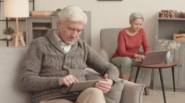 Orta boylu, beyaz bir adam koltukta oturuyor, tablet bilgisayar kullanıyor, sonra koltukta kısa saçlı Asyalı bir kadınla konuşuyor, dizüstü bilgisayarla arka planda yazı yazıyor.