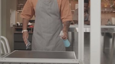 Sabahleyin açılmadan önce kahve dükkanında önlük giyen ve masaları silen kısa saçlı, çift ırklı bir kadın, arka planda tezgahın arkasında duran siyah bir erkek arkadaş.