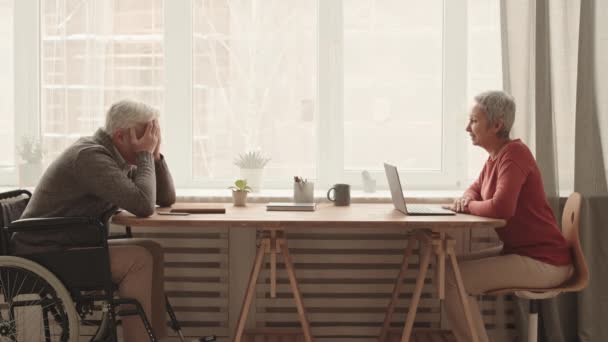白发苍苍的亚洲女人白天坐在窗前 与坐在轮椅对面的沮丧的白人老人交谈 手挽手 — 图库视频影像