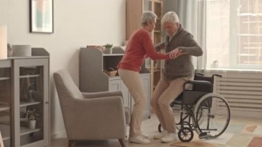 Kısa saçlı, Asyalı bir kadının, yaşlı beyaz bir adamın tekerlekli sandalyeden kalkmasına ve gündüz vakti oturma odasında koltuğa oturmasına yardım edişinin tam kenarı.