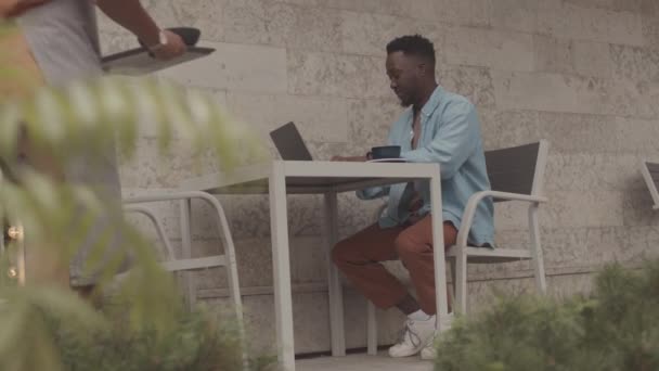 用便携式计算机向坐在室外餐桌旁的年轻黑人男子提供盘中餐的低角度小吃 — 图库视频影像