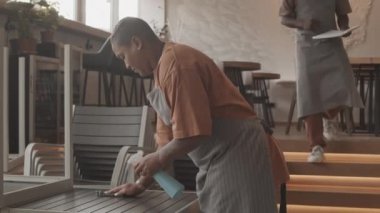 Sabah açılmadan önce kahve dükkanında kısa saçlı melez bir kadının önlük giyip, temizlik masasını silmesi, arka planda yürüyen siyahi bir erkek arkadaşın...