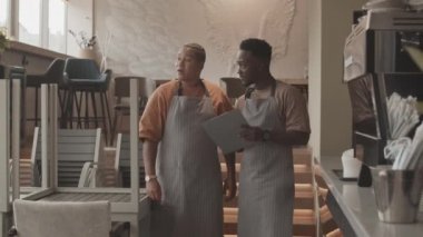 Orta uzunlukta, önlük giymiş genç bir çift ırklı kadın ve elinde pano olan siyah bir adam kahve dükkanında dikilip vardiyadan önce görevlerini tartışıyor.