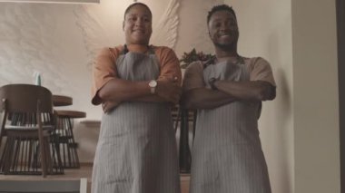 Düşük açılı orta ölçekli genç siyahi erkek ve melez kadının önlük giyip, restoranda dikilip kollarını göğsüne dolayıp kameraya bakıp gülümsediği bir sahne.