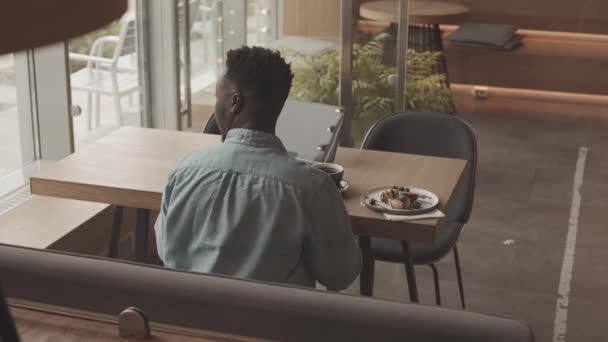 黑人男子的后排高角形 坐在餐馆餐桌前 吃早餐 用手提电脑 裁剪过的侍者拿着无线支付终端走近他 客户用智能手机付款 — 图库视频影像