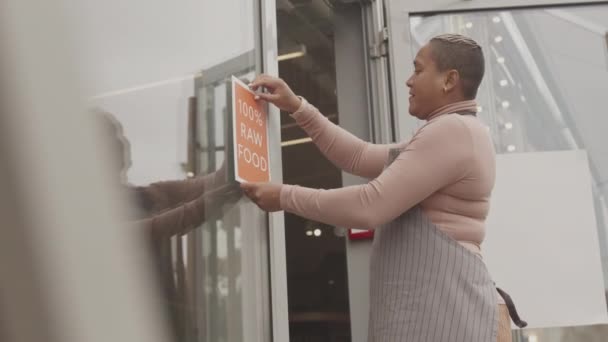 エプロンを身に着けている若い産生の女性の所有者の低角度中長 店外の入り口に100 生の食品記号を置く — ストック動画