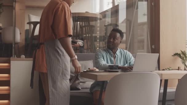 中くらいの若いです黒男のテーブルでレストランで座って ポータブルコンピュータを使用して クロップされたBiracialウェイターは クライアントからの支払いを得るために無線支払い端末で彼に近づいて — ストック動画
