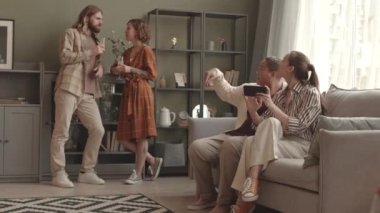 Beyaz bir kadın ve erkek oturma odasında oturmuş, köpüklü şarap içip sohbet ederken, iki bayan arkadaş kanepede oturmuş, akıllı telefondan video izliyorlar.