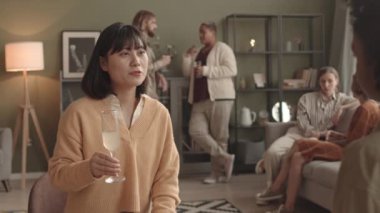 Orta boy Asyalı bir kadın, gündüz vakti oturma odasında oturmuş, kırpılmış arkadaşlarıyla kokteyl içiyor, bardakları tokuşturuyor, konuşuyor ve gülümsüyor, arka planda bulanık insanlar takılıyor.