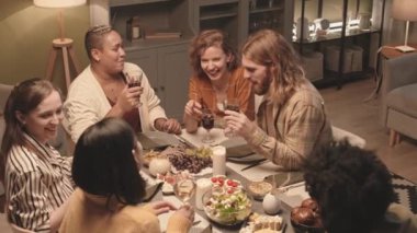 Altı farklı etnik gruptan genç arkadaşın evde yemek masasında oturup sohbet etmesi, şarap kadehlerini kaldırması, şapırdatması ve gülümsemesi.