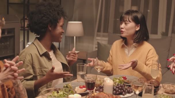 非洲裔美国和亚裔年轻妇女在夜间坐在客厅的餐桌前 进行令人兴奋的交谈 饮酒和庆祝活动 — 图库视频影像