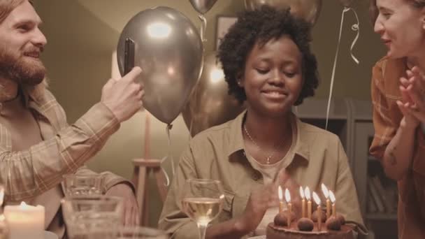 若いアフリカ系アメリカ人の誕生日の女の子の下で チョコレートケーキにろうそくを吹き消し 願い事をし 白人女性は手を拍手し 男はスマートフォンで撮影し そしてすべての巨大な — ストック動画