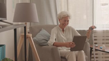 Dijital tablette sohbet eden neşeli yaşlı kadın videosunun orta ölçekli fotoğrafı rahat bir dairede, yumuşak koltukta oturuyor.