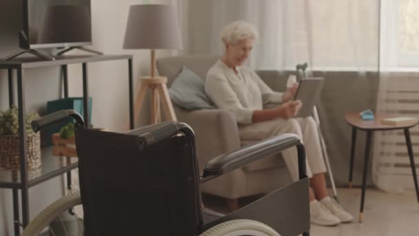 一个心不在焉的老年妇女坐在家里与医生在线咨询 在平板电脑上通过视频通话 讨论药物和药物的镜头 轮椅站在前排 — 图库视频影像