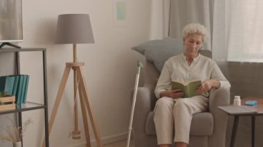 Orta boy, kır saçlı, kitap okuyan, rahat bir apartmanın penceresinde, yumuşak koltukta oturan yaşlı kadının fotoğrafı.